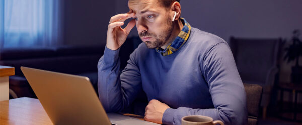 A man with short hair sits at a desk, looking intently at a laptop screen while resting his forehead on his hand. He wears a blue sweater with a collared shirt that has a checkered pattern. A beige mug and a smartphone are on the table in front of him, and the room is dimly lit.