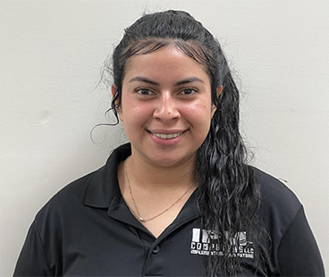 A young woman with long, wavy black hair is smiling at the camera, wearing a black polo shirt with the logo of a company on the left side. The background is plain and light-colored.