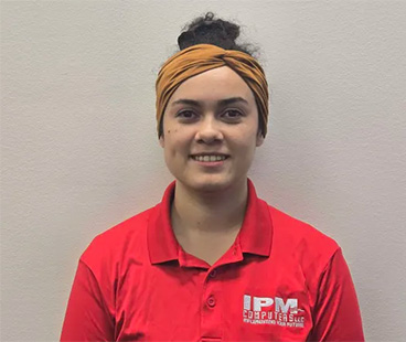 Marisa Flournoy A person with dark hair styled in a bun, wearing an orange headband and a red polo shirt with the logo "IPM Computers LLC," smiles in front of a neutral-colored wall.