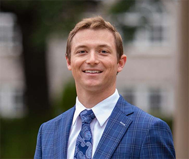 Peyton Whisnant A smiling young man in a blue checkered suit with a white shirt and patterned tie stands outdoors, with blurred buildings and greenery in the background.