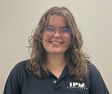 Taylor Moore A young woman with curly hair and glasses smiles at the camera, wearing a dark polo shirt with the letters "IPM" on it. The background is a plain light color.