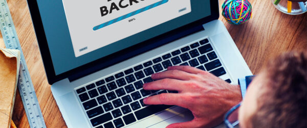 Data Backup in Progress A person’s hand typing on a laptop displaying a "DATA BACKUP" progress bar at 100%. A cup of coffee is placed beside the laptop on a wooden desk.