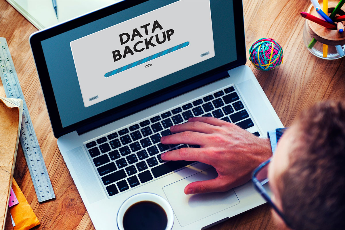 A person’s hand typing on a laptop displaying a "DATA BACKUP" progress bar at 100%. A cup of coffee is placed beside the laptop on a wooden desk.