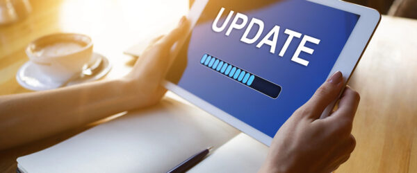 Device Update A person holding a tablet displaying a blue screen with the word "UPDATE" and a loading progress bar, with a notebook and a cup of coffee in the background.