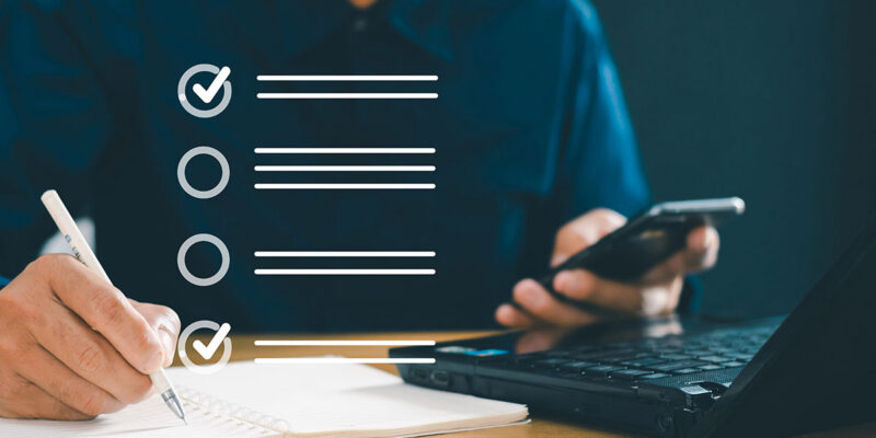 Compliance Checklist A person writing in a notebook with a pen while holding a smartphone, next to a laptop. Three checklist items are illustrated, with one item marked as complete.