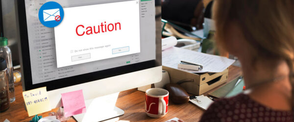 A person sitting at a desk using a computer, with an email window displaying a red caution message on the screen. The workspace is cluttered with papers, sticky notes, and a coffee mug.