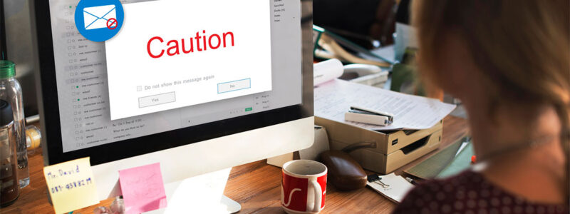 A person sitting at a desk using a computer, with an email window displaying a red caution message on the screen. The workspace is cluttered with papers, sticky notes, and a coffee mug.