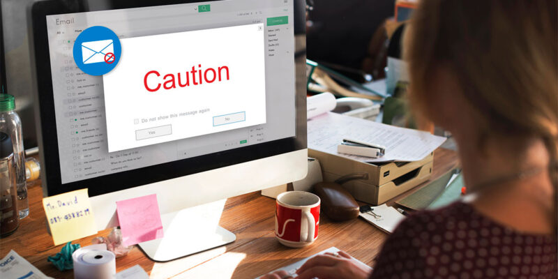 A person sitting at a desk using a computer, with an email window displaying a red caution message on the screen. The workspace is cluttered with papers, sticky notes, and a coffee mug.