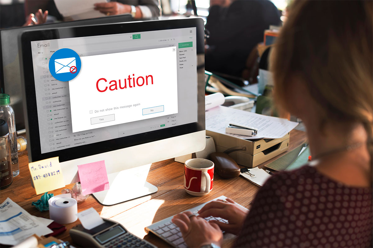 A person sitting at a desk using a computer, with an email window displaying a red caution message on the screen. The workspace is cluttered with papers, sticky notes, and a coffee mug.