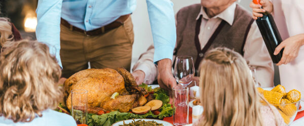 A festive dining table with a roasted turkey at the center, surrounded by various side dishes. A man in a blue shirt serves the turkey while an elderly man in a sweater looks on. Children with curly hair and a girl with long hair sit at the table, watching the scene. A woman pours wine from a bottle.