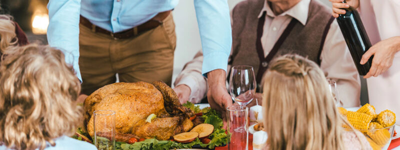 A festive dining table with a roasted turkey at the center, surrounded by various side dishes. A man in a blue shirt serves the turkey while an elderly man in a sweater looks on. Children with curly hair and a girl with long hair sit at the table, watching the scene. A woman pours wine from a bottle.