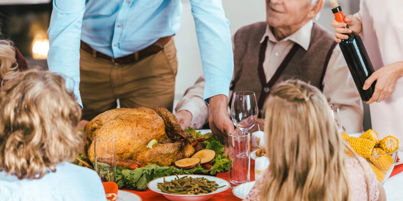 A festive dining table with a roasted turkey at the center, surrounded by various side dishes. A man in a blue shirt serves the turkey while an elderly man in a sweater looks on. Children with curly hair and a girl with long hair sit at the table, watching the scene. A woman pours wine from a bottle.