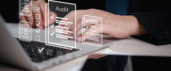 A close-up of hands typing on a laptop with digital audit checklists and documents displayed above the keyboard.