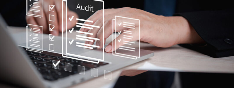 A close-up of hands typing on a laptop with digital audit checklists and documents displayed above the keyboard.
