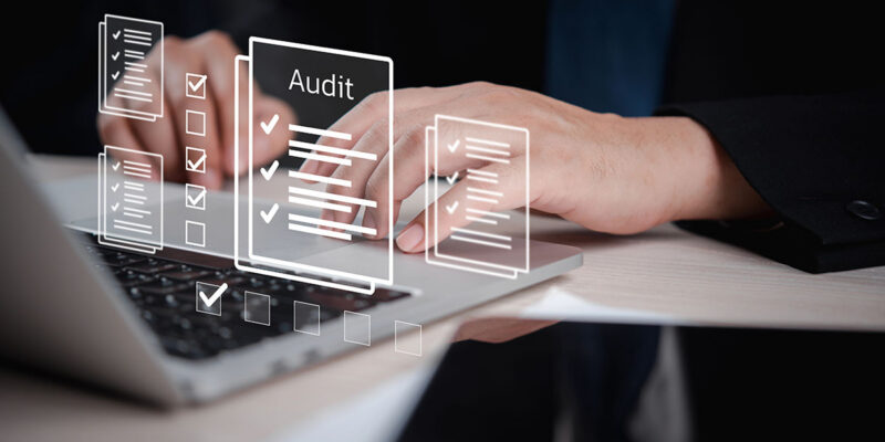 A close-up of hands typing on a laptop with digital audit checklists and documents displayed above the keyboard.