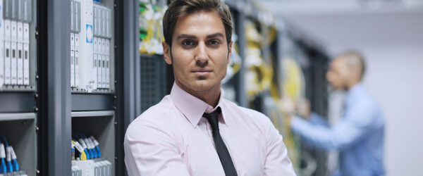 A confident man in a pink dress shirt and black tie stands with his arms crossed in front of server racks in a data center, while another professional in a blue shirt works in the background.