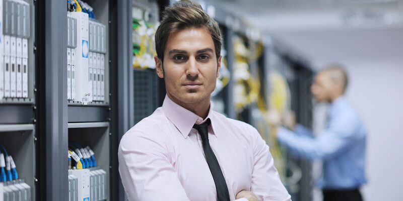 A confident man in a pink dress shirt and black tie stands with his arms crossed in front of server racks in a data center, while another professional in a blue shirt works in the background.