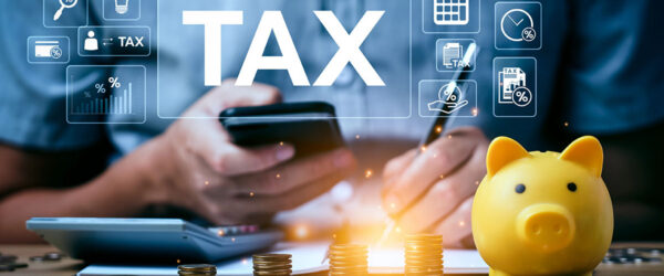 A person holds a smartphone while using a pen to write on a document near a calculator, coins, and a yellow piggy bank; digital icons related to taxes are displayed above.