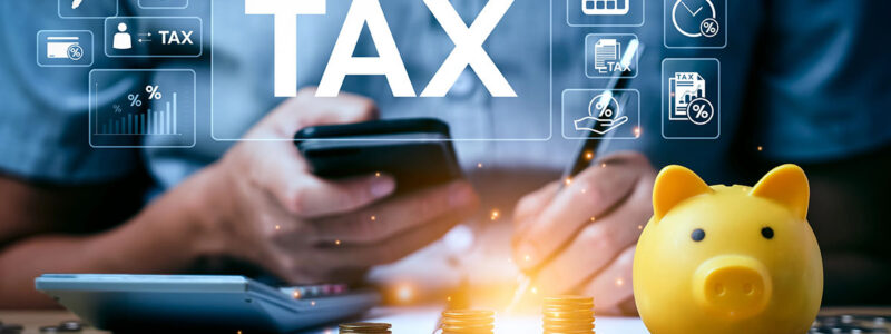 A person holds a smartphone while using a pen to write on a document near a calculator, coins, and a yellow piggy bank; digital icons related to taxes are displayed above.