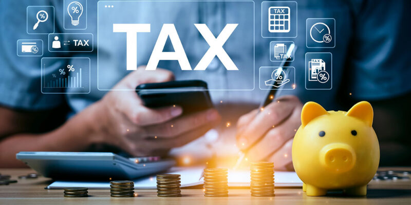 A person holds a smartphone while using a pen to write on a document near a calculator, coins, and a yellow piggy bank; digital icons related to taxes are displayed above.