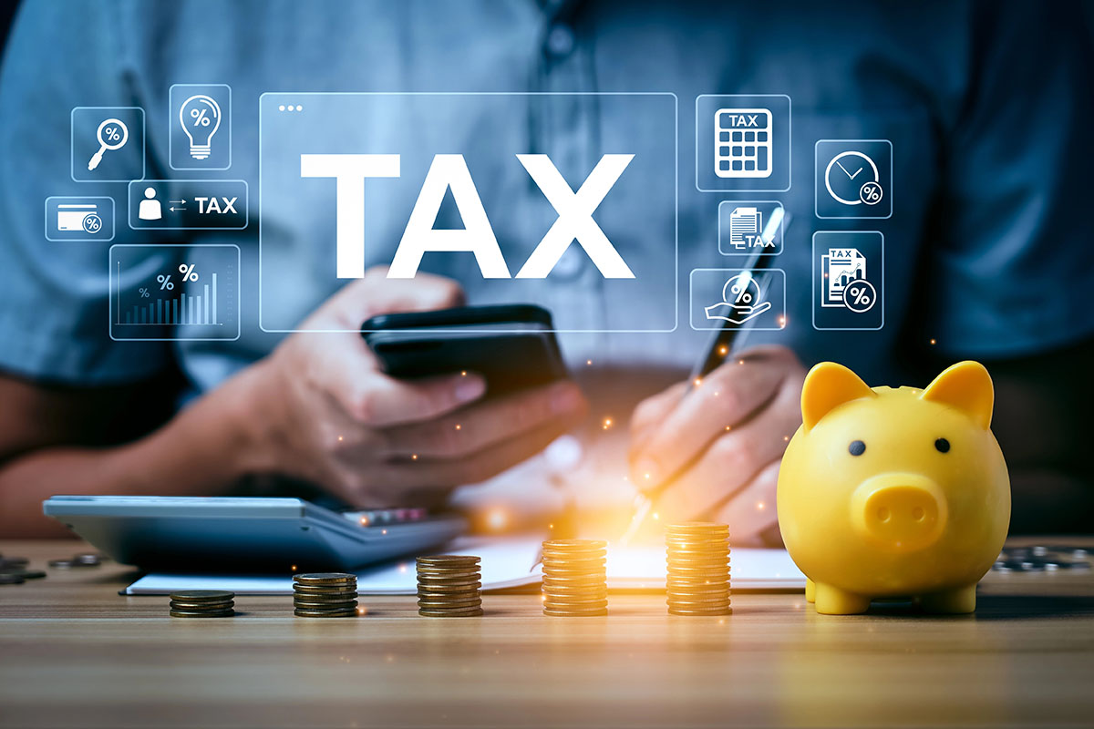 A person holds a smartphone while using a pen to write on a document near a calculator, coins, and a yellow piggy bank; digital icons related to taxes are displayed above.