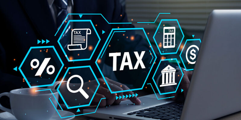A businessman in a suit is using a laptop with digital tax-related icons, including documents, percentage symbols, and a bank, overlaid in a futuristic graphic style. A cup of coffee and glasses are placed on the table.