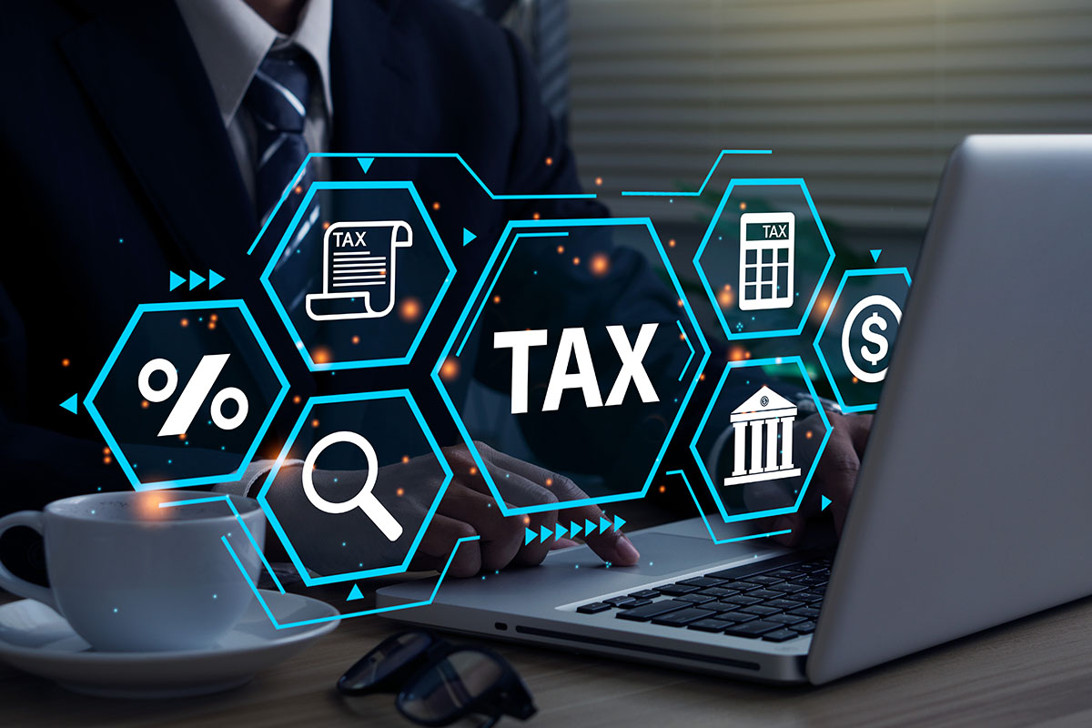A businessman in a suit is using a laptop with digital tax-related icons, including documents, percentage symbols, and a bank, overlaid in a futuristic graphic style. A cup of coffee and glasses are placed on the table.