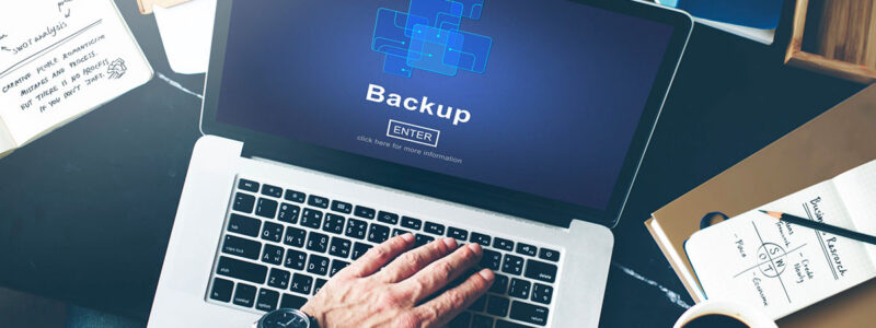 A laptop displaying a "Backup" screen with an "ENTER" prompt, with a person's hand hovering over the keyboard. A notepad and coffee cup are visible on a desk with various stationary items.