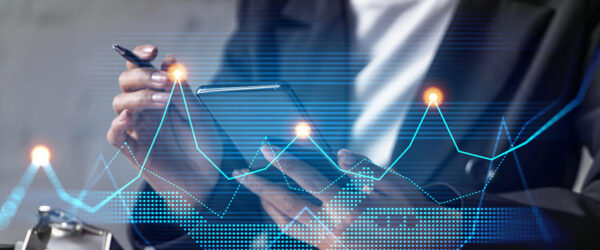 A person in a business suit is holding a smartphone in one hand and a pen in the other, with digital graphs and data visualizations overlaying the scene.