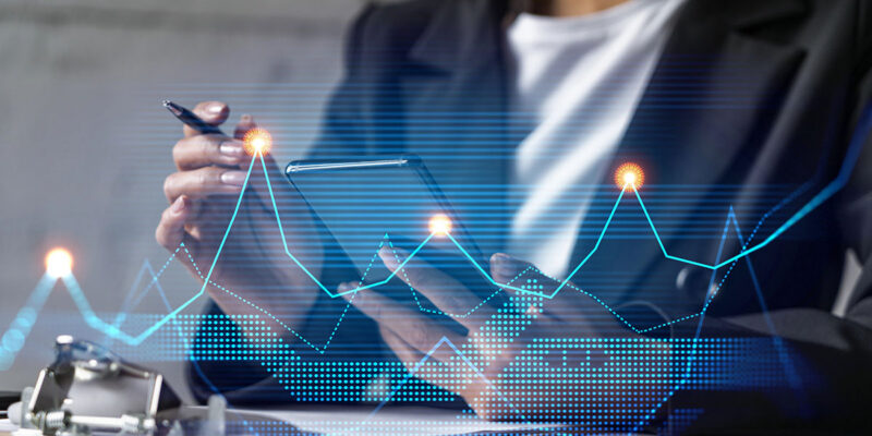 A person in a business suit is holding a smartphone in one hand and a pen in the other, with digital graphs and data visualizations overlaying the scene.