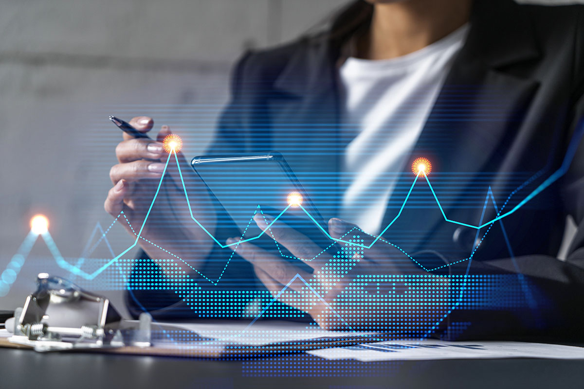 A person in a business suit is holding a smartphone in one hand and a pen in the other, with digital graphs and data visualizations overlaying the scene.