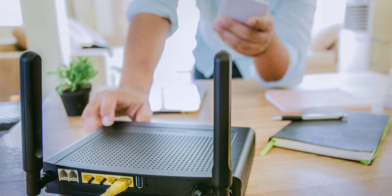 A person adjusts a black router on a wooden table, with a yellow Ethernet cable connected to it. In one hand, they hold a smartphone, while notebooks and a small plant are visible in the background.