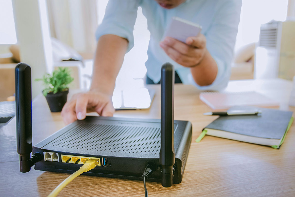 A person adjusts a black router on a wooden table, with a yellow Ethernet cable connected to it. In one hand, they hold a smartphone, while notebooks and a small plant are visible in the background.
