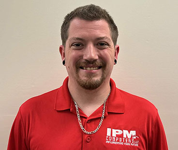 Austin B. A smiling man with short, light brown hair and ear piercings is wearing a red polo shirt with the logo "IPM COMPUTERS, LLC." against a plain background.