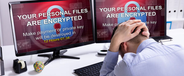 A man sits at a desk with his head down, appearing stressed. Two computer monitors display a warning message: "YOUR PERSONAL FILES ARE ENCRYPTED" and a countdown timer, indicating 12 hours and 1 minute remaining to make a payment or risk losing files. Office supplies are visible on the desk.