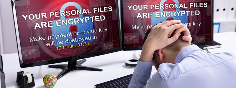 A man sits at a desk with his head down, appearing stressed. Two computer monitors display a warning message: "YOUR PERSONAL FILES ARE ENCRYPTED" and a countdown timer, indicating 12 hours and 1 minute remaining to make a payment or risk losing files. Office supplies are visible on the desk.