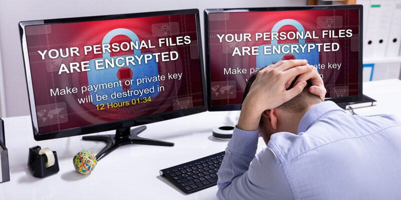 A man sits at a desk with his head down, appearing stressed. Two computer monitors display a warning message: "YOUR PERSONAL FILES ARE ENCRYPTED" and a countdown timer, indicating 12 hours and 1 minute remaining to make a payment or risk losing files. Office supplies are visible on the desk.
