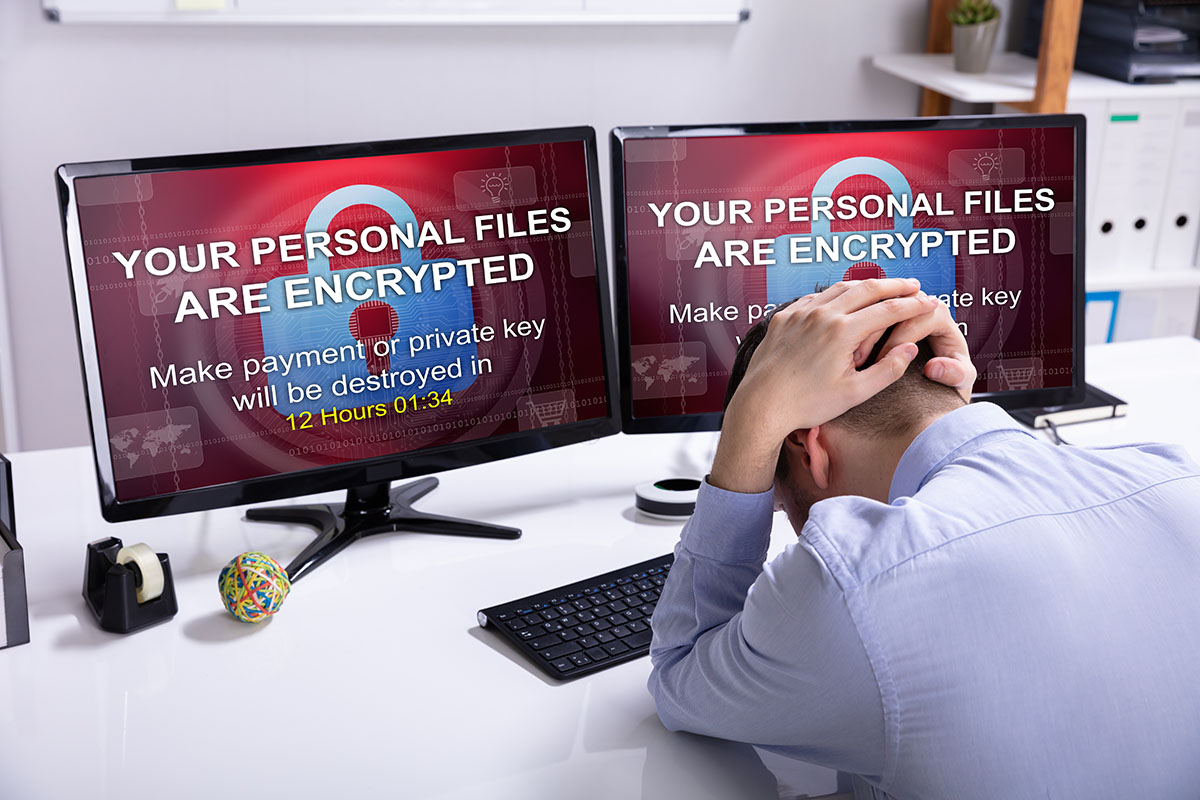 A man sits at a desk with his head down, appearing stressed. Two computer monitors display a warning message: "YOUR PERSONAL FILES ARE ENCRYPTED" and a countdown timer, indicating 12 hours and 1 minute remaining to make a payment or risk losing files. Office supplies are visible on the desk.