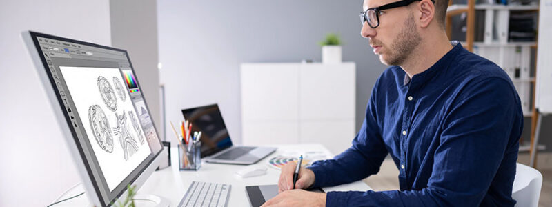 A man with glasses is sitting at a modern desk, working on a digital tablet while looking at a large computer monitor displaying graphic designs. Color swatches are arranged on the desk, and a small plant is visible in the background.