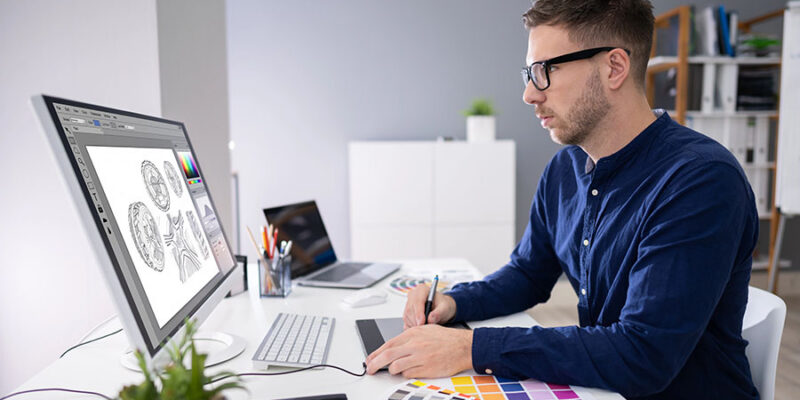 A man with glasses is sitting at a modern desk, working on a digital tablet while looking at a large computer monitor displaying graphic designs. Color swatches are arranged on the desk, and a small plant is visible in the background.