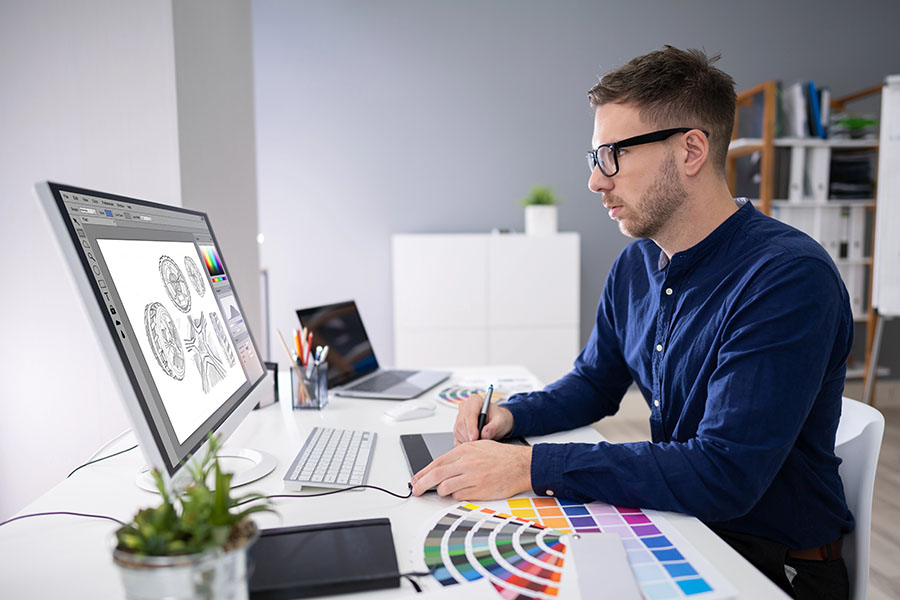 A man with glasses is sitting at a modern desk, working on a digital tablet while looking at a large computer monitor displaying graphic designs. Color swatches are arranged on the desk, and a small plant is visible in the background.
