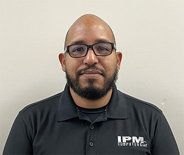 A man with a beard and glasses is smiling, wearing a black polo shirt with the logo "IPM COMPUTER LLC," against a plain light-colored background.