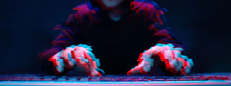 A hooded figure sits in front of a keyboard in a dark setting, with hands hovering over the keys, creating an abstract, glitch-like effect in vibrant blue and red tones.