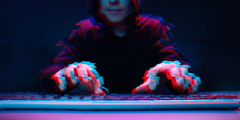 A hooded figure sits in front of a keyboard in a dark setting, with hands hovering over the keys, creating an abstract, glitch-like effect in vibrant blue and red tones.