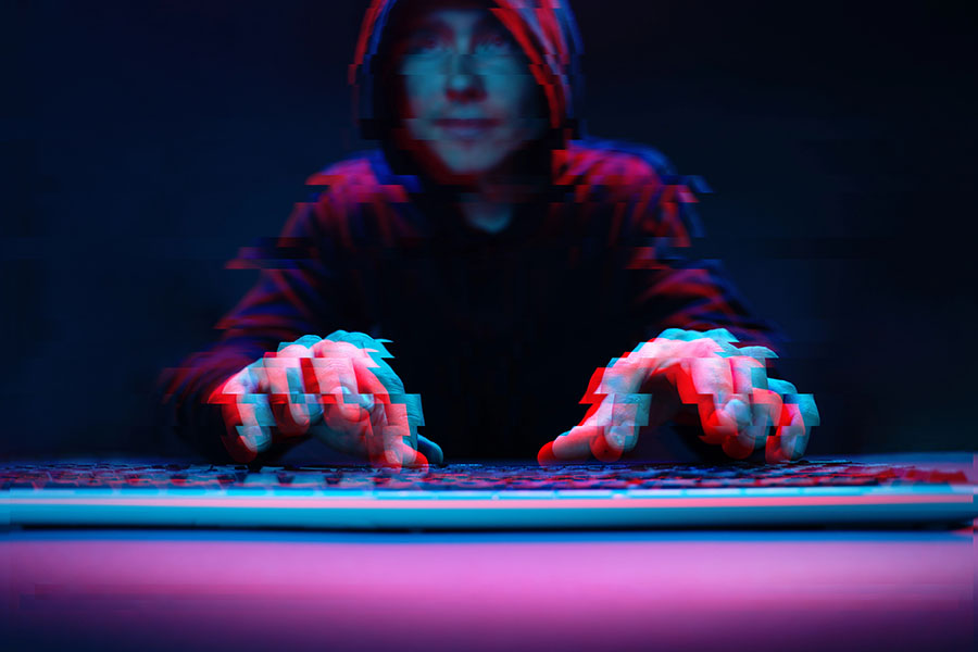 A hooded figure sits in front of a keyboard in a dark setting, with hands hovering over the keys, creating an abstract, glitch-like effect in vibrant blue and red tones.