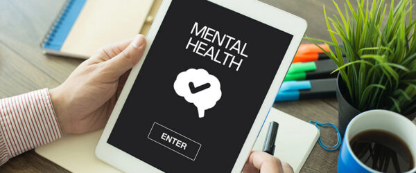 A person holding a tablet displaying the words "MENTAL HEALTH," with a graphic of a brain and a check mark above a button labeled "ENTER." The background includes a notebook, colorful pens, and a coffee cup on a wooden table.