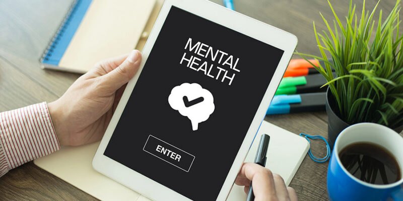 A person holding a tablet displaying the words "MENTAL HEALTH," with a graphic of a brain and a check mark above a button labeled "ENTER." The background includes a notebook, colorful pens, and a coffee cup on a wooden table.
