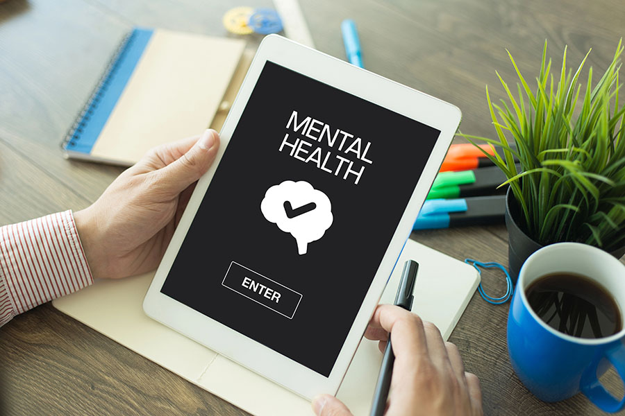 A person holding a tablet displaying the words "MENTAL HEALTH," with a graphic of a brain and a check mark above a button labeled "ENTER." The background includes a notebook, colorful pens, and a coffee cup on a wooden table.