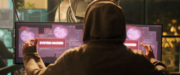 A person in a hoodie viewed from behind, gesturing triumphantly in front of dual computer monitors displaying red "SYSTEM HACKED" warning pop-ups.