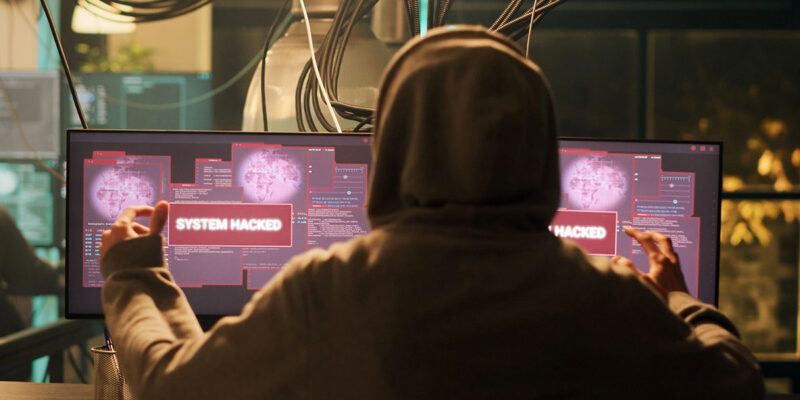 A person in a hoodie viewed from behind, gesturing triumphantly in front of dual computer monitors displaying red "SYSTEM HACKED" warning pop-ups.